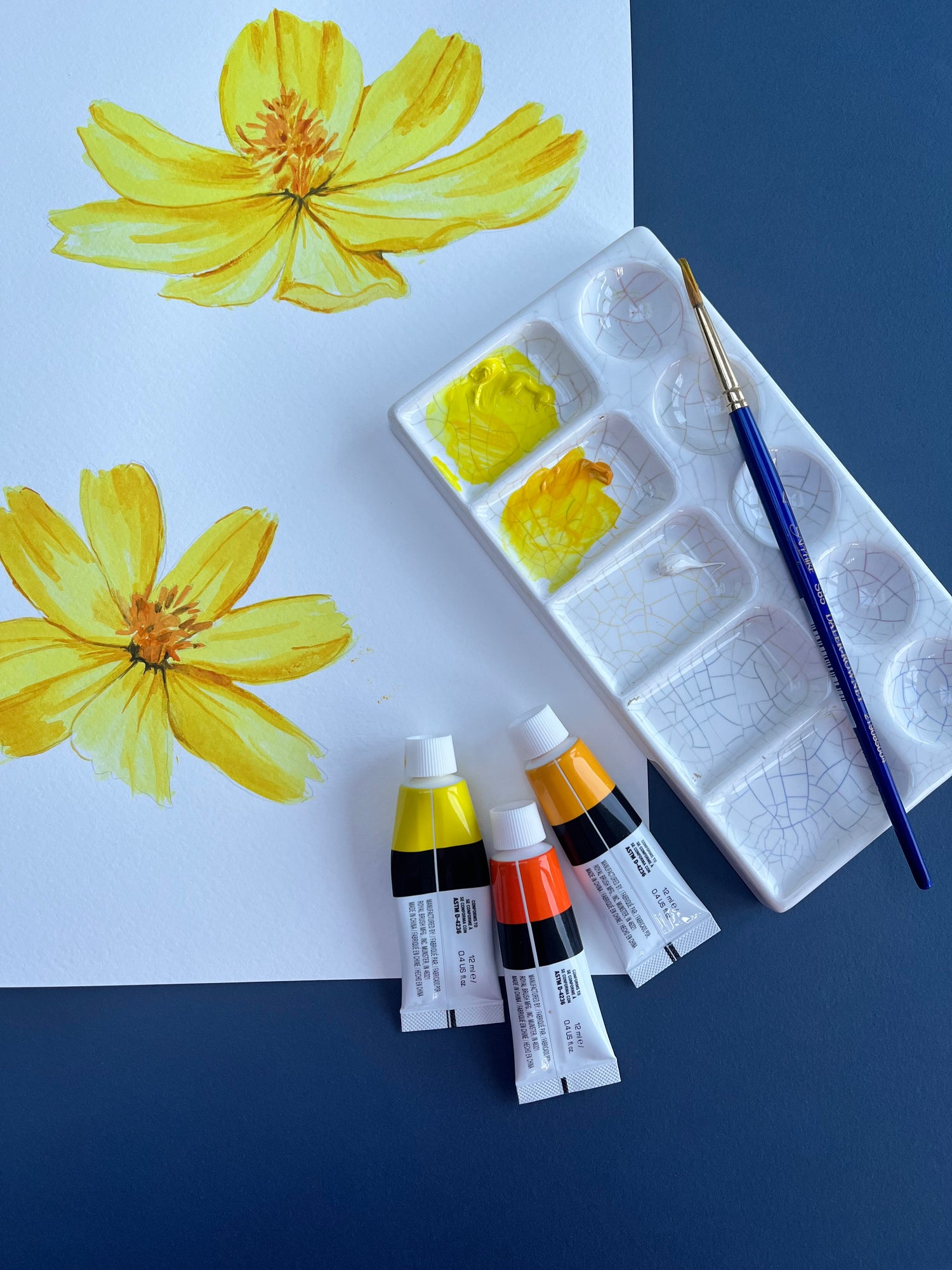 Art supplies including a paint palette, tubes of paint, and a brush on a paper with yellow flower paintings.