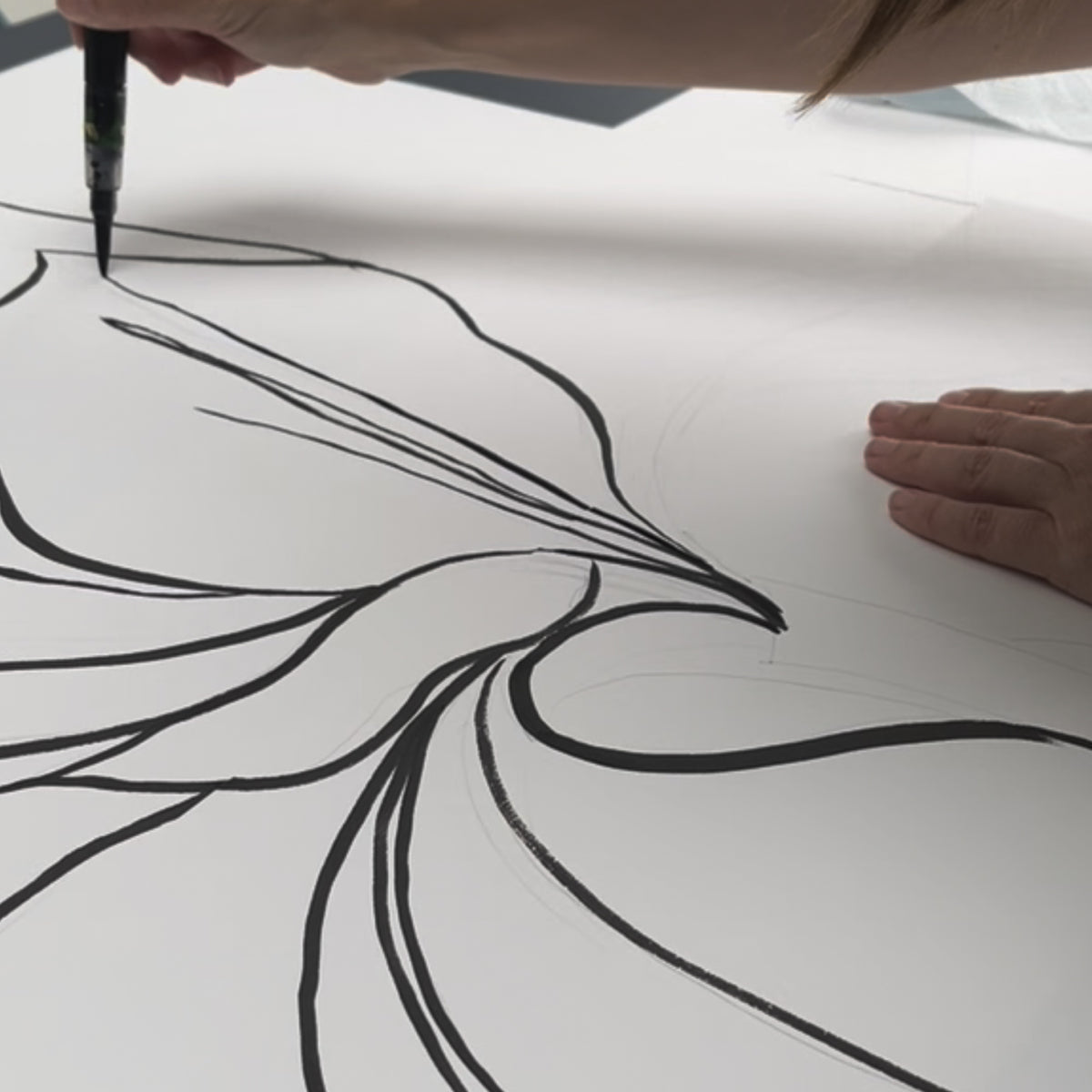 A close-up of a Sarah's hand holding a black brush pen, drawing a bold black line for the Iris wall art on a large white sheet of paper. Another hand rests on the right side of the paper.
