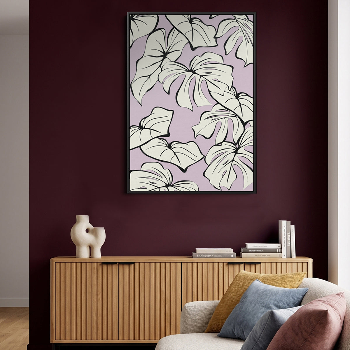 Framed Kai line art wall print featuring large white monstera leaves on a light lavender background, hanging on a deep burgundy wall above a light wood console table in a styled living room.