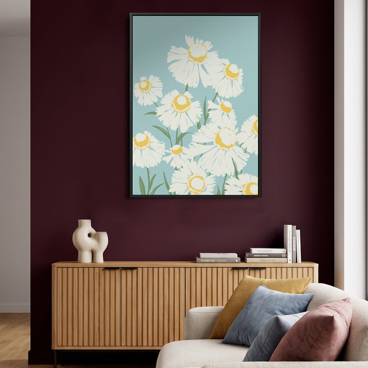 The Margarite Light Blue framed wall art print featuring white daisies with yellow centres on a light blue background, hung on a dark burgundy wall above a wooden console table in a styled living room.